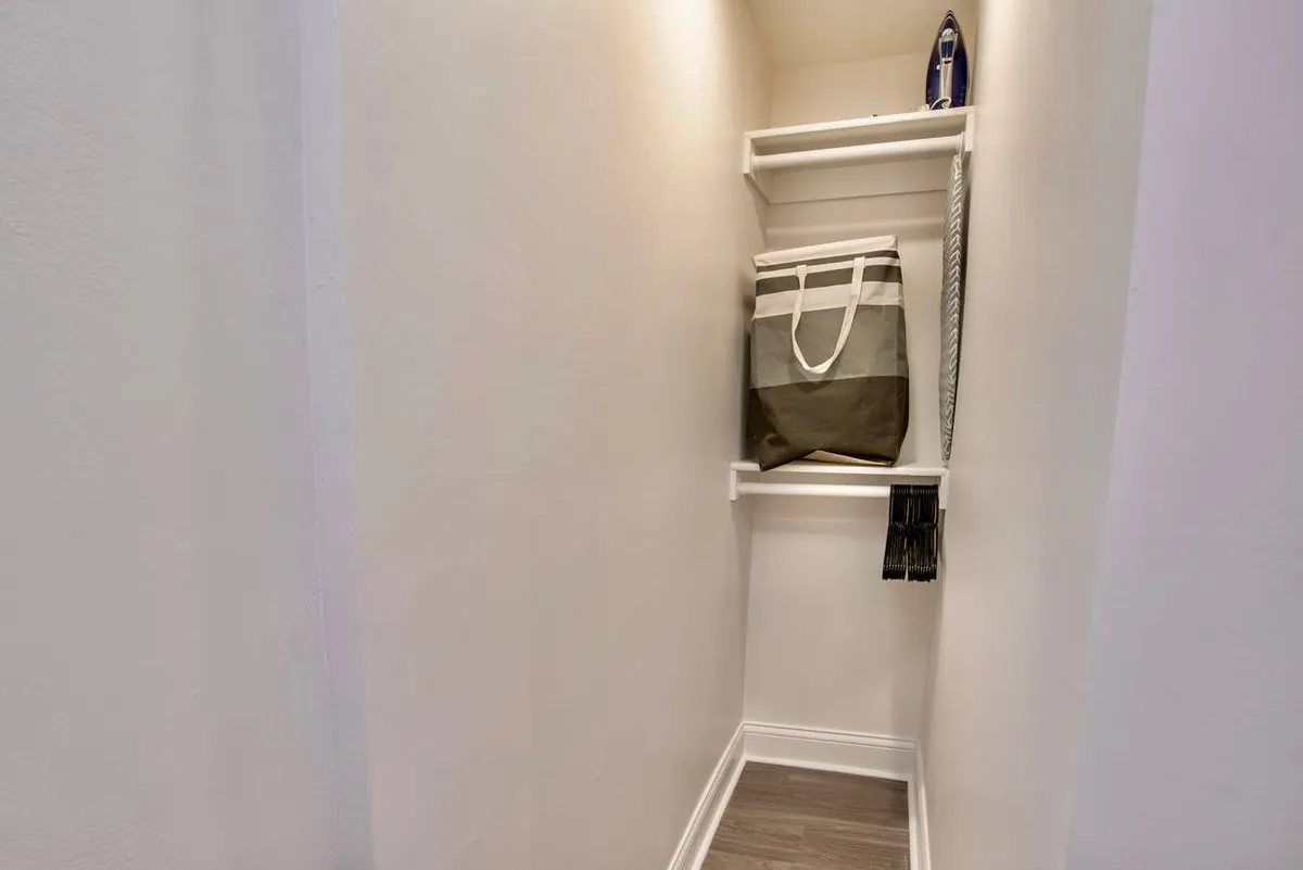 A functional storage space within the apartment, featuring open shelves, a bag, and an iron. The area is tidy and organized, suggesting thoughtful amenities for guests’ practical needs.