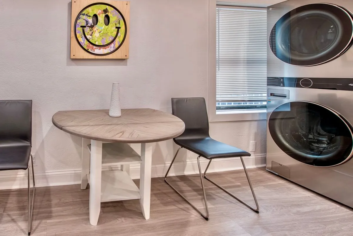 This image presents a cheerful dining area with a round table and modern chairs, set against playful artwork and a relaxed atmosphere. The table is inviting, ideal for casual meals, conversations, or gathering with friends. The inclusion of a laundry machine subtly hints at the multi-functional nature of the home without distracting from the dining focus.