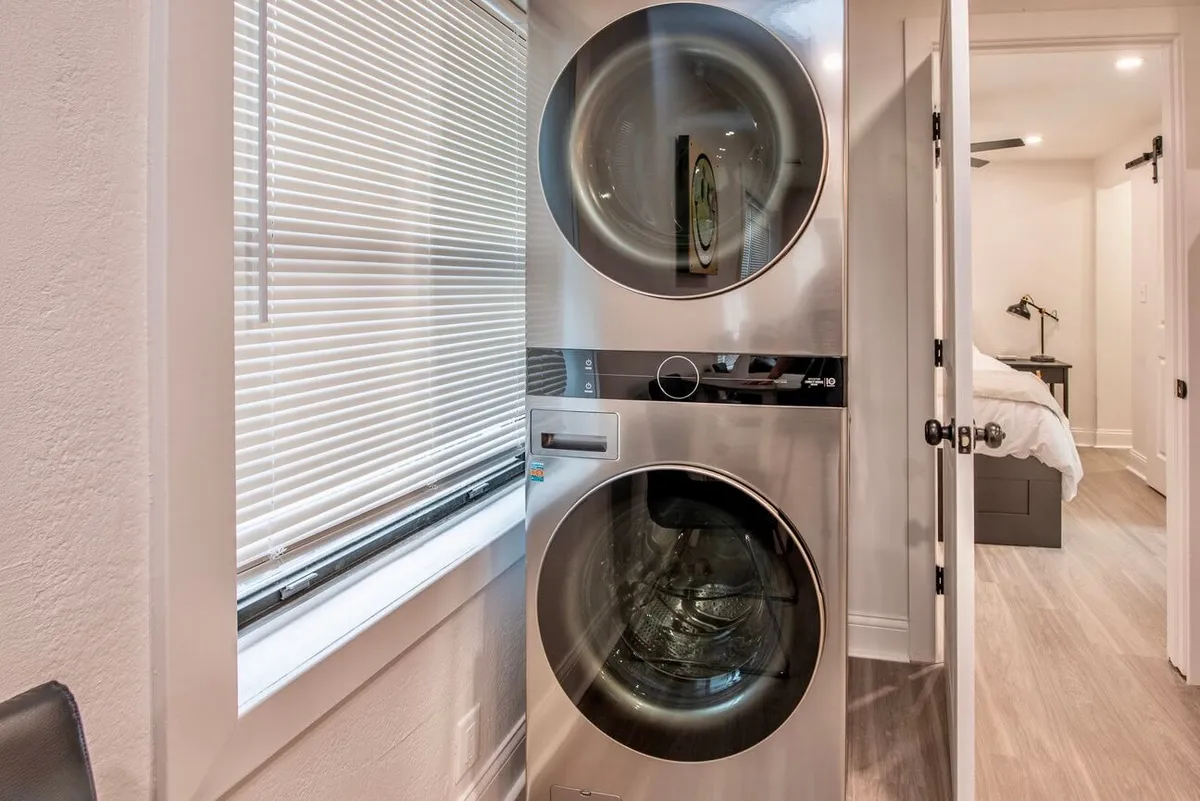 A view of a contemporary apartment’s laundry area, showcasing a stacked washer and dryer next to a window with blinds. The space is bright and neatly organized, conveying a sense of modern efficiency.
