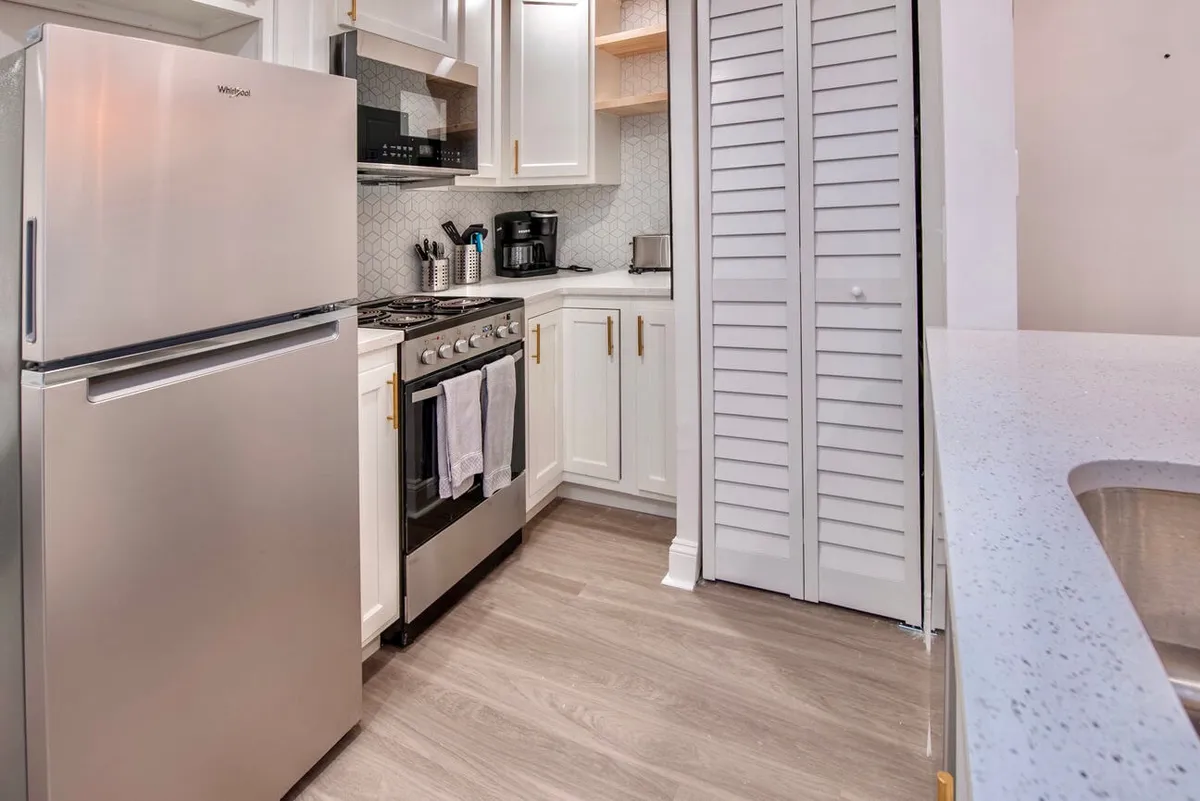 This image showcases a modern kitchen equipped with a stainless steel refrigerator and gas stove, complemented by sleek white cabinetry and a granite countertop. A coffee maker sits on the counter, hinting at both daily rituals and the potential for entertaining. The space is bright and inviting, with ample room for meal preparation and gathering.