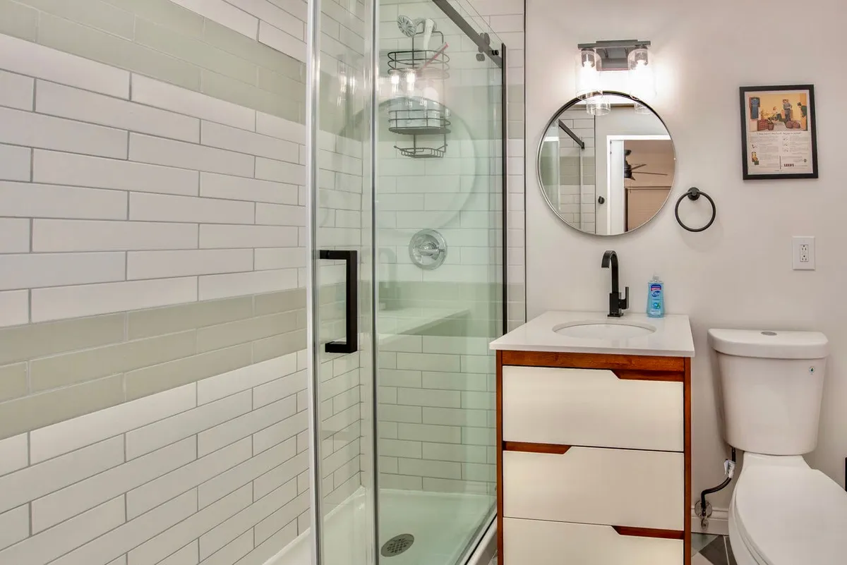 A modern bathroom with a glass-door shower, a vanity accented with natural wood, a stylish round mirror, and contemporary light fixtures. The space feels fresh and clean, ideal for both invigorating morning or relaxing evening routines.